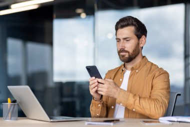 Modern bir ofis masasında çalışan genç profesyonel telekomünikasyon, akıllı telefonla mesajlaşırken dizüstü bilgisayara odaklanma, iş ve uzaktan çalışma için çağdaş bir çalışma alanında birden fazla iş yapma