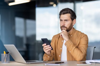 Tecrübeli işadamı ofis masasında oturuyor, cep telefonunu tutuyor, iş sırasında düşünüyor, karmaşık görevler için çözümler buluyor veya yeni fikirler üretiyor.