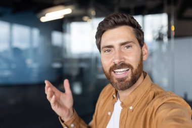 Gülümseyen sakallı adam kameraya birinci şahıs gözüyle bakarken el hareketi yapıyor. Modern ofis ortamında kendine güvenini gösteriyor.