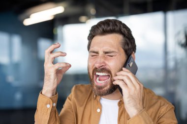 Stresli bir adam ofisteki akıllı telefonunda zor bir konuşma yaparken gözleri kapalı bağırıyor ve el hareketleriyle tedirgin oluyor.
