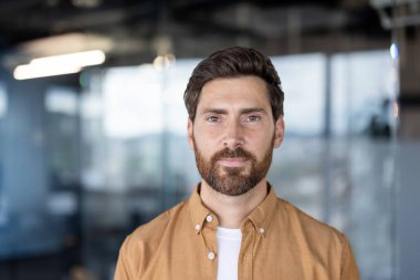 Kendine güvenen sakallı bir adam, bulanık modern ofis ortamında dikilirken kameraya doğrudan bakıyor. İş ortamında profesyonelliği, başarıyı ve liderliği simgeliyor.