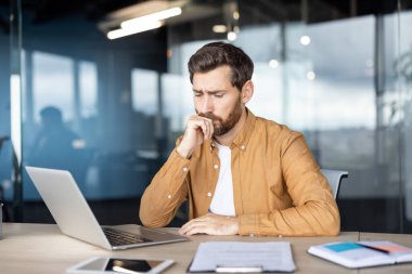 Ofis masasında oturan işadamı, sorunlu ve bilgisayarına odaklanmış bir halde, bir problem ya da yeni bir proje hakkında derin bir şekilde düşünüyor, kendini kararsız hissediyor ve iş sırasında konsantre oluyor.