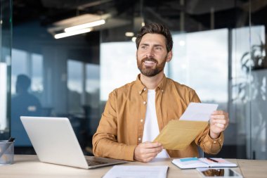 Ofis masasındaki mutlu işadamı posta zarfından çıkarılan bir belgeyi okurken gülümsüyor, olumlu yazışmaları, onayları veya başarılı iş haberlerini kutluyor.