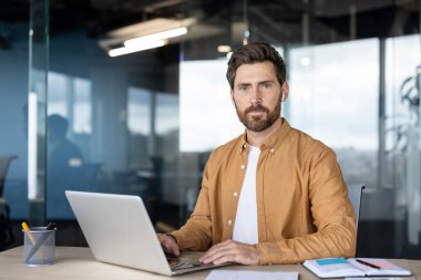 Sakallı ve kablosuz kulaklıklı bir adam laptopuyla meşgul, çağdaş ofis ortamında bir masada oturuyor, odaklanmış çalışmayı ve yenilikçi teknoloji kullanımını temsil ediyor.