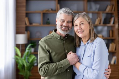 Üst düzey karı ve kocayı sevmek, el ele tutuşmak ve sarılmak, modern bir oturma odasında kameraya bakarken mutluluk ve sadakat ifade etmek.