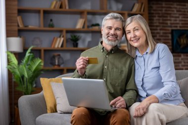 Olgun bir çift kameraya gülümsüyor, evdeki koltukta oturuyor, elinde kredi kartı olan bir adam online alışveriş için dizüstü bilgisayar kullanıyor, modern teknolojiyi kucaklıyor.