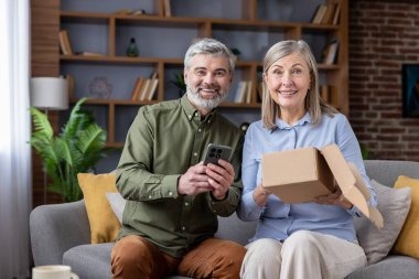 Evdeki kanepede oturan mutlu yaşlı çift. Kadın paketini açarken erkek akıllı telefonunda online alışveriş ya da teslimat onayı arıyor.