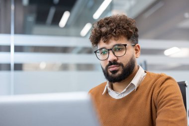 Kıvırcık saçlı, sakallı ve gözlüklü genç profesyonel bir adam modern bir ofiste dizüstü bilgisayarla çalışıyor, strateji, verimlilik ve dijital gelişime odaklanıyor.