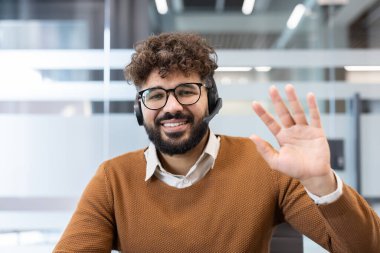 Ofisinden uzak bir video konferansında kameraya el sallayan, gözlüklü, kıvırcık saçlı gülümseyen bir adam profesyonel müşteri desteği ve sanal işbirliği teklif ediyor.