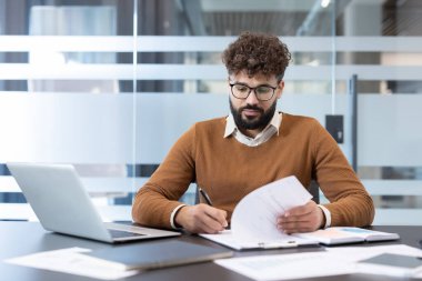 Sakallı, kıvırcık saçlı, gözlüklü, belgelere yazmaya odaklanmış, şirket ortamında dizüstü bilgisayarlı bir masada otururken evrakları inceleyen genç bir adam.