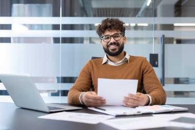 Kameraya bakan ve elinde kağıtlarla mutlu bir işadamı, modern bir ofis masasında bir dizüstü bilgisayar ve çeşitli belgelerle çalışıyor, başarı ve profesyonelliği temsil ediyor.