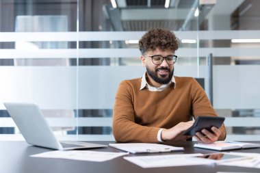 Finansal verileri analiz eden ve hesap makinesiyle hesaplamalar yapan genç bir adam modern bir ofiste masasında çalışırken, işletme yönetimi ve muhasebe üzerine odaklanır.