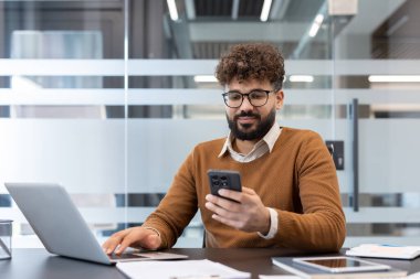 Modern bir ofiste çalışan genç profesyonel bir adam bilgisayarlı ve belgeli bir masada otururken akıllı telefonuna bakıyor. İletişim ve çoklu görevi temsil ediyor.
