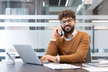 Gözlüklü profesyonel bir adam telefonda konuşurken kameraya gülümser ve modern bir ofiste dizüstü bilgisayarda yazar. Yoğun bir iş gününde birden fazla iş yapar.