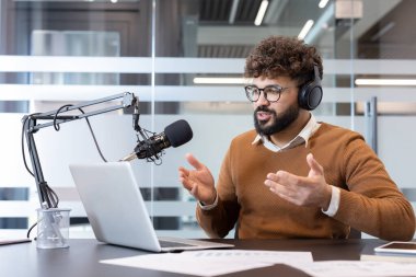 Gözlüklü ve kulaklıklı genç adam podcast kaydediyor ya da profesyonel bir mikrofona canlı yayın yapıyor, bir dizüstü bilgisayarda çalışıyor ve modern bir ofis masasında belgeler hazırlıyor.