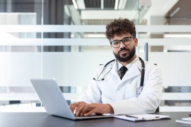 Steteskoplu genç erkek doktor beyaz laboratuvar önlüğü ve gözlüğü giyiyor, dizüstü bilgisayarla yazarken kameraya bakıyor, modern bir klinikte ya da hastane ofisinde çalışıyor, sağlık hizmetleri sunuyor.