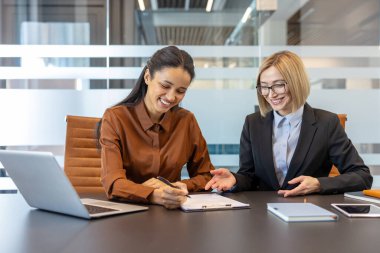İki gülümseyen iş kadını, modern bir ofis ortamında karanlık bir konferans masasında oturmuş, bir panoda iş birliği yapıyor ve önemli bir anlaşmayı imzalıyor.