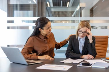 Stresli bir meslektaşı destekleyen ve rahatlatan iş kadını, modern ofis ortamında baş ağrısı ve tükenme, empati ve takım çalışması sergiliyor.