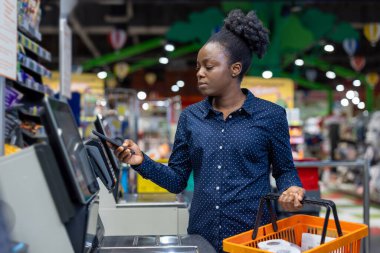 Elinde akıllı telefon tutan genç bir kadın, modern bir süpermarkette otomatik kontrol makinesine bağlanmadan ödeme yapıyor, kontrol sürecine kolaylık ve verimlilik getiriyor.
