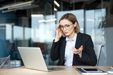 Stresli iş kadını kafasına dokunuyor. Modern bir şirket ofisinde bilgisayarında çalışırken baş ağrısı ve hayal kırıklığı belirtileri gösteriyor.