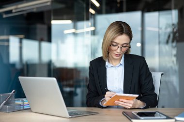 Günümüz ofis ortamında dizüstü bilgisayarlı bir masada çalışırken, gözlük takan genç bir iş kadını, bir dizüstü bilgisayarla çalışırken, organizasyon, odak ve verimlilik sunuyor.