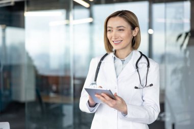 Beyaz laboratuvar önlüğü ve steteskop giyen kadın doktor, dijital tablet ile konuşurken gülümsüyor, modern sağlık hizmetleri iletişimini ve teknolojisini temsil ediyor.