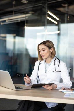 Beyaz önlüklü ve steteskoplu kadın doktor bir masada oturuyor, elinde kalem ve tablet, sanal bir tıbbi danışma sırasında dizüstü bilgisayarı işaret ediyor veya çevrimiçi bir konferans sırasında