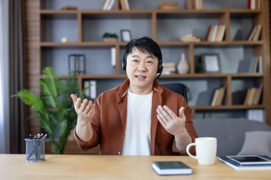Kahverengi tişörtlü ve kulaklıklı Asyalı bir adam internet üzerinden yaptığı bir video görüşmesi sırasında ya da e-öğrenim oturumunda konuşuyor ya da uzaktan destek veriyor ya da öğretiyor.
