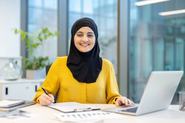 Sarı tişörtlü, tesettürlü Müslüman bir kadın masasında çalışırken kameraya bakıp gülümsüyor, dizüstü bilgisayar kullanıyor ve modern bir ofisteki bir panoya not yazıyor.