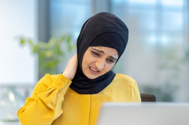 Modern ofis ortamında dizüstü bilgisayarı üzerinde çalışırken başörtüsü takan Müslüman kadın, zayıf ergonomik ve işle ilgili stresle ilgili sorunları vurguluyor.