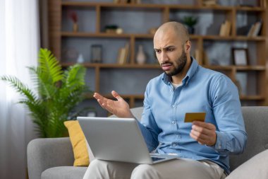 Evde dizüstü bilgisayar kullanarak online alışveriş yapan, kredi kartı işlemleri veya ödemeleriyle ilgili sorunları olan ve ödemelerde düşüş yaşanmasına yol açan sinirli bir adam.