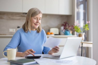 Üst düzey kadın, modern mutfağında dizüstü bilgisayar ve hesap makinesi kullanırken kişisel finans ve ev faturalarını özenle kontrol ediyor.