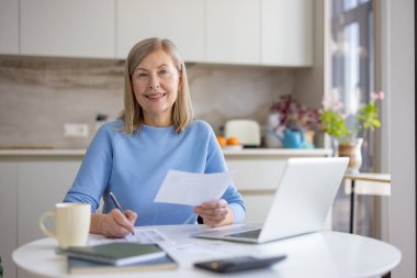 Üst düzey bir kadın mutfak masasında ev ekonomisini incelerken gülümsüyor. Faturaları ödemek için dizüstü bilgisayar, hesap makinesi ve evrakları kullanıyor.