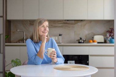 Beyaz bir mutfak masasında oturan, elinde bir kupayla düşünceli bir şekilde yüzünü çeviren, evinde sakin ve mutlu bir an geçiren son sınıf öğrencisi kadın.