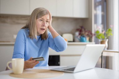 Üst düzey kadın stresli ve kafası karışık görünüyor, evde dizüstü bilgisayarın önünde otururken elinde bir kredi kartı tutuyor, internette dolandırıcılık ya da siber suç dolandırıcılığı potansiyeli var.