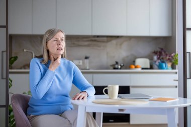 Boğazı ağrıyan ve acı çeken yaşlı bir kadın, mutfak masasında oturup boynuna dokunuyor, evde otururken rahatsızlık ve hastalık yaşıyor.