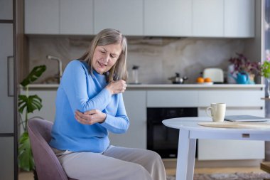 Üst düzey bir kadın modern bir mutfak masasında oturuyor, iki eliyle dirseğini buruyor ve sıkıyor, eklem ağrısı ve eklem iltihabı ya da evde yaralanma yüzünden acı çekiyor.