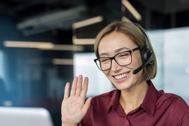 Kulaklık ve reçeteli gözlük takan genç bir kadın, sanal bir video görüşmesi sırasında elini sallarken mutlu bir şekilde gülümsüyor, ofis ortamından dostane müşteri hizmetleri ve destek sağlıyor.