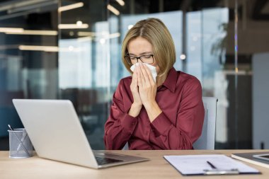 Modern bir ofiste soğuk algınlığı ya da alerjik reaksiyon gösteren bir iş kadını masasında dizüstü bilgisayarla otururken burnunu mendile sümkürdü.