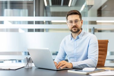 Masasında dizüstü bilgisayarda çalışan, iş hayatında başarı ve teknoloji kullanımını temsil eden çağdaş ofis ortamında kameraya bakan, kendine güvenen erkek girişimci.