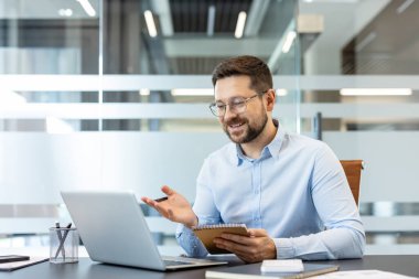 İş adamı, modern bir ofisten laptopuyla bir video görüşmesine aktif olarak katılıyor, notlar alıyor ve sanal bir toplantı sırasında fikirleri tartışıyor.