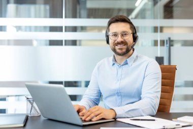 Modern bir ofiste çalışan gülümseyen bir adam, mikrofonlu bir kulaklık takıyor, online müşteri hizmetleri sağlıyor ve bir dizüstü bilgisayar kullanırken teknik destek sağlıyor.