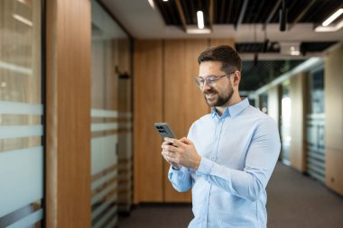 Modern bir ofis koridorunda duran genç işadamı, iletişim ve ağ kurmak için akıllı telefonu güvenle kullanıyor, profesyonel bir iş ortamında teknoloji entegrasyonunu yansıtıyor