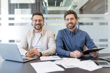 Çağdaş bir ofiste masa başında birlikte oturan iki neşeli erkek profesyonel dizüstü bilgisayar üzerinde çalışırken piyasa raporlarını ve belgelerini gözden geçiriyorlar, takım çalışmasını somutlaştırıyorlar ve başarılı bir ortaklık kuruyorlar.