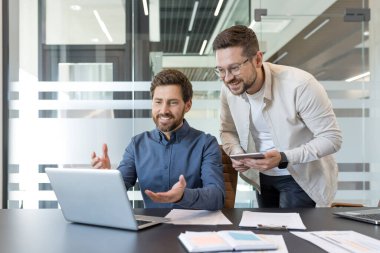 İki gülümseyen erkek profesyonel, modern bir dizüstü bilgisayar ve tablet üzerinde çalışarak çağdaş bir iş yerinde başarılı takım çalışmalarını teşvik ederek verimli bir tartışmaya girişiyor.