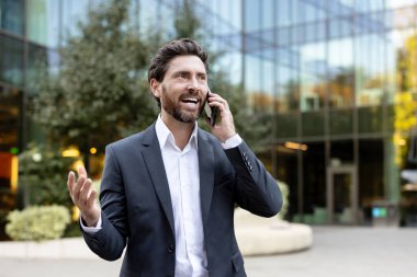Dışarıda dikilen ve telefonda konuşan mutlu iş adamı, zarif iş kıyafetleri içinde iletişim kuruyor ve neşe veriyor.