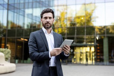 Kendine güvenen sakallı iş adamı modern cam ofis binasının önünde duruyor, dijital tabletle tutuyor ve etkileşime geçiyor, profesyonel hayatta teknoloji entegrasyonunu temsil ediyor.