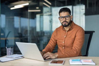 Masasında oturan, dizüstü bilgisayarda yazan ve modern ofis ortamında kameraya bakan, üretkenliği ve profesyonelliği temsil eden genç ve ciddi bir iş adamı.