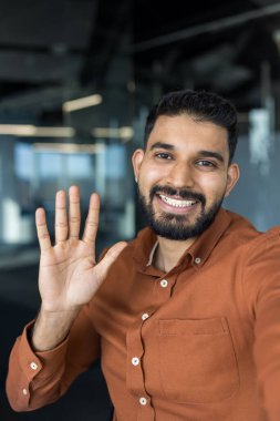 Dost canlısı genç bir adam kameraya bakıyor ve elini sallıyor, çevrimiçi bir toplantı sırasında selamlıyor ya da veda ediyor ya da çağdaş bir ofis ortamında neşeli bir selfie çekiyor.