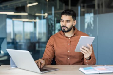 Genç adam işe odaklanmış, dizüstü bilgisayar kullanıyor ve modern bir ofiste dijital tablet tutuyor, kurumsal bir ortamda yenilik, bağlantı ve verimli online iletişimi sembolize ediyor.